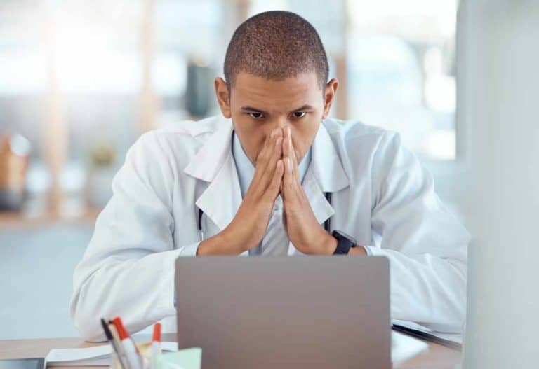 A doctor sitting at his desk with his hands on his face, exhausted from dealing with the aftermath of a ransomware attack in his clinic's cybersecurity systems.
